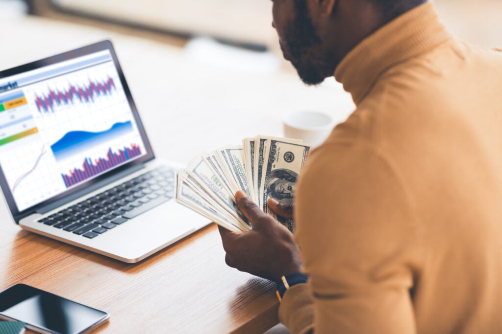 Person holding cash while looking at financial data on a laptop in a bright workspace.